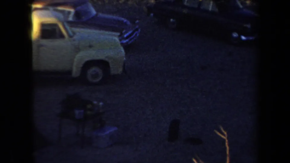 Low-light Arizona camping frame with a cropped pickup, dark sedan, small gear, and gravel surface.