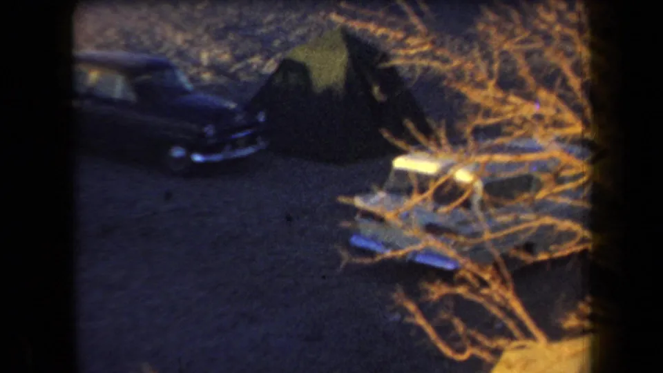 1958 Arizona 8mm frame showing a dim parked car near dry brush and a tent-like shape at a nighttime campsite.