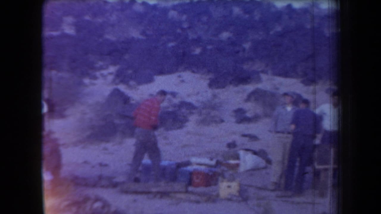 Adult men in 1958 Arizona appear gathered around camp gear on a rocky hillside in soft, purple-tinted 8mm footage.