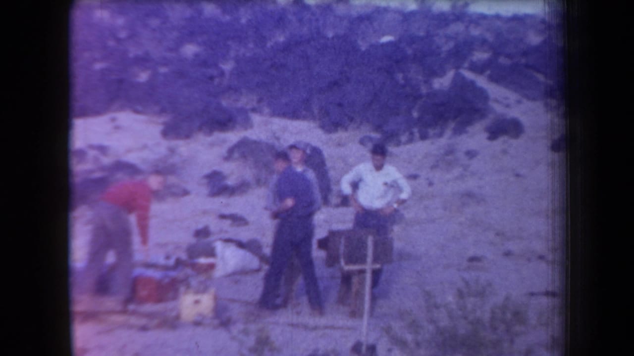 Several men stand near camp gear and a small frame or chair in rocky terrain.