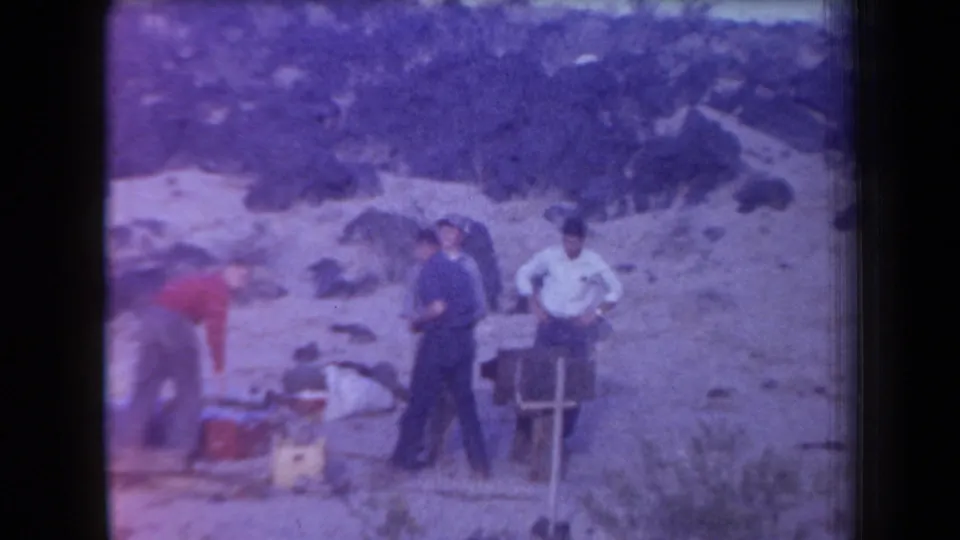 Several adult men stand near coolers, boxes, and a small chair or frame on an Arizona rocky hillside.