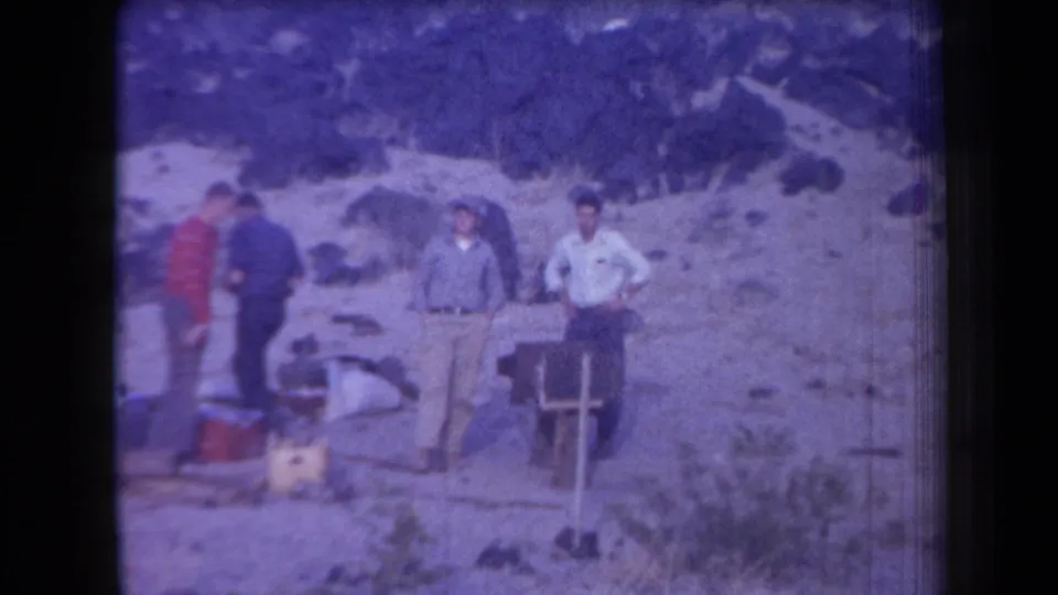 Four adult men in casual shirts and trousers stand by coolers and gear in a rocky Arizona outdoor setting.