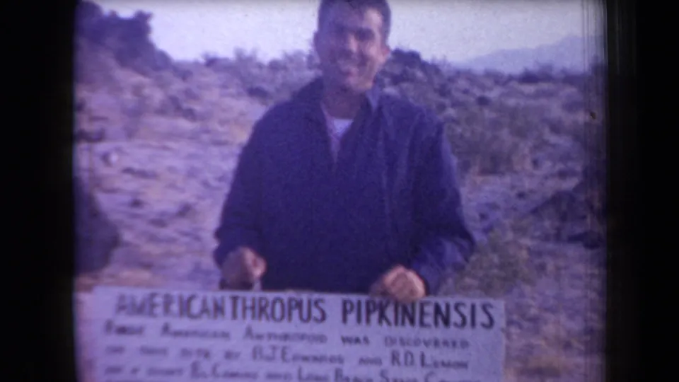 A man in 1958 Arizona holds a fossil discovery sign in front of rocky desert terrain, with the sign text partly legible.