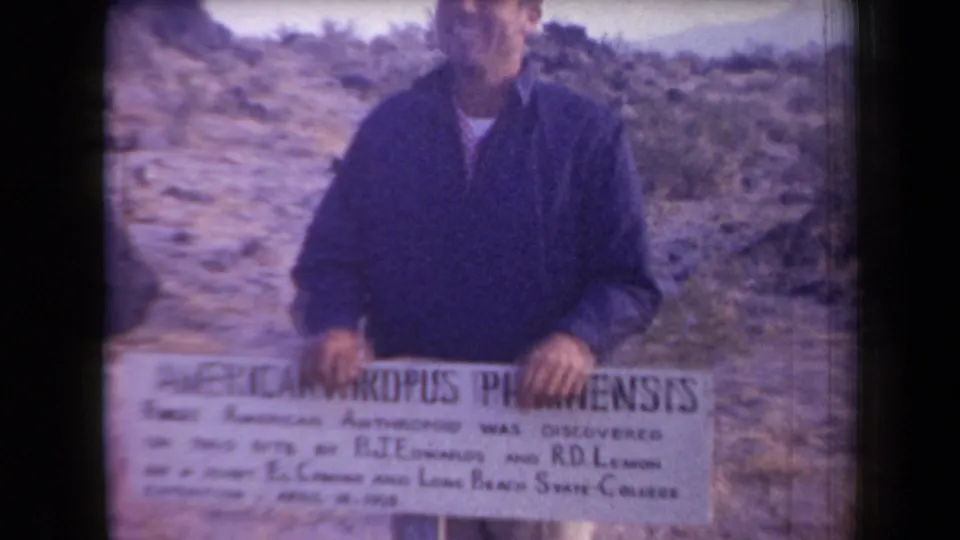 In 1958 Arizona home-movie footage, a man faces the camera behind a sign reading AMERICANTHROPUS PIPKINENSIS in a rocky desert landscape.