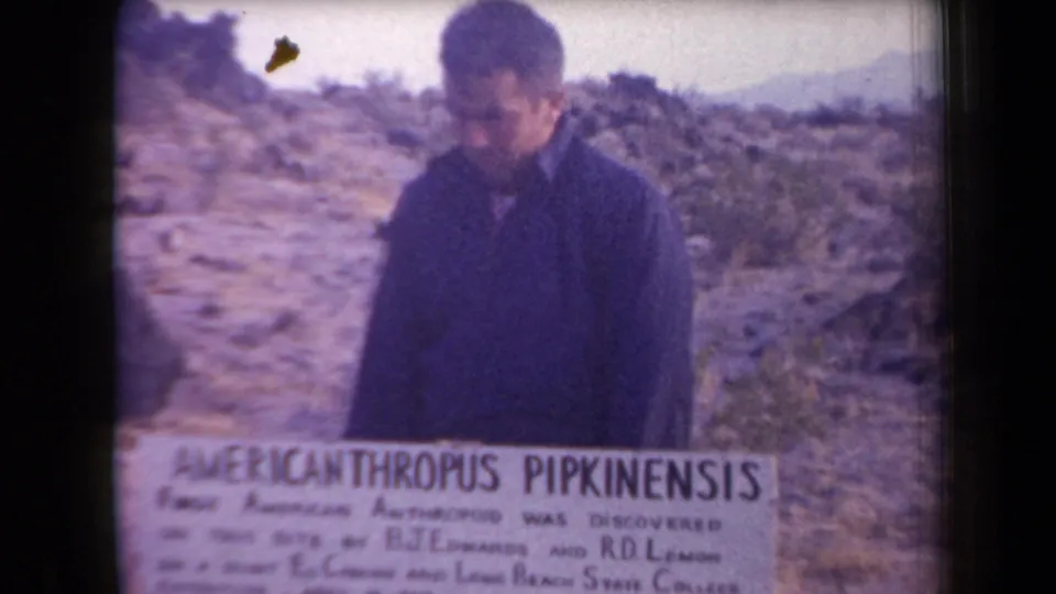 In a soft 8mm frame, the man lowers his gaze behind the fossil discovery sign amid rocky Arizona desert terrain.