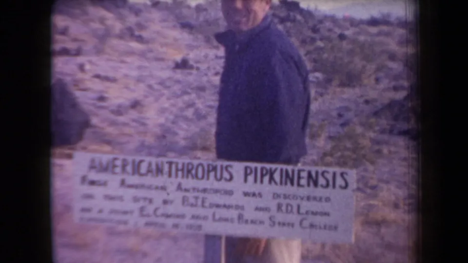 The man turns partly sideways beside the AMERICANTHROPUS PIPKINENSIS sign, with arid hills and sparse vegetation behind him.