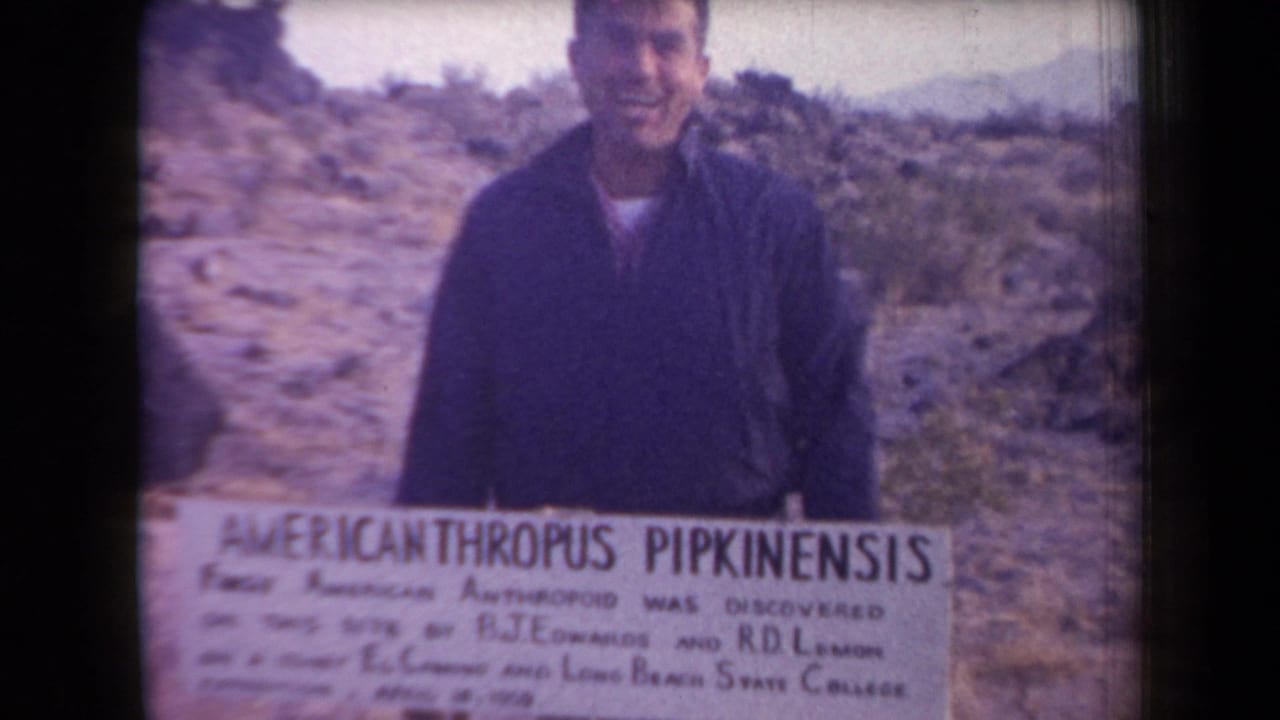 In 1958 Arizona 8mm footage, an adult man smiles behind a white humorous sign in a rocky desert landscape.