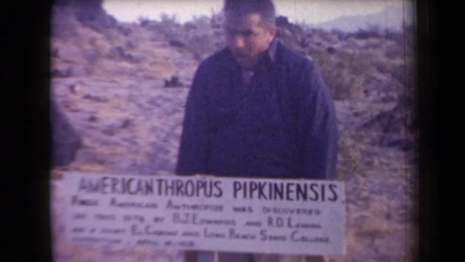 The man stands close to the readable novelty sign, looking downward amid Arizona desert rocks and scrub.