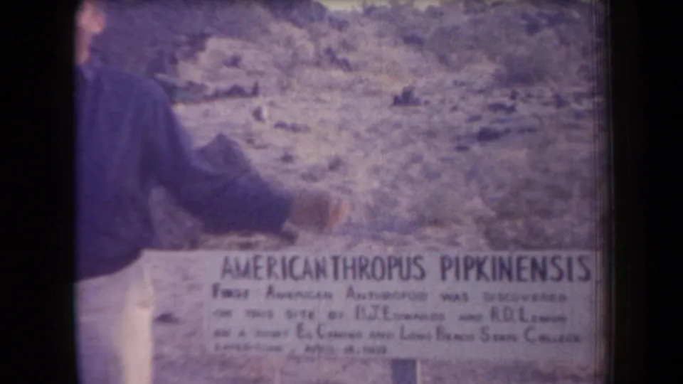 A cropped view shows the man reaching toward the Americanthropus Pipkinensis sign against rocky Arizona desert terrain.
