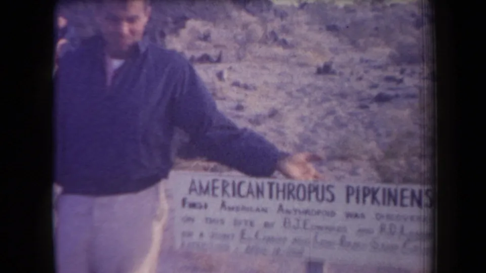 The man stands beside the novelty sign and gestures toward it, with scrub-covered desert and hills behind him.