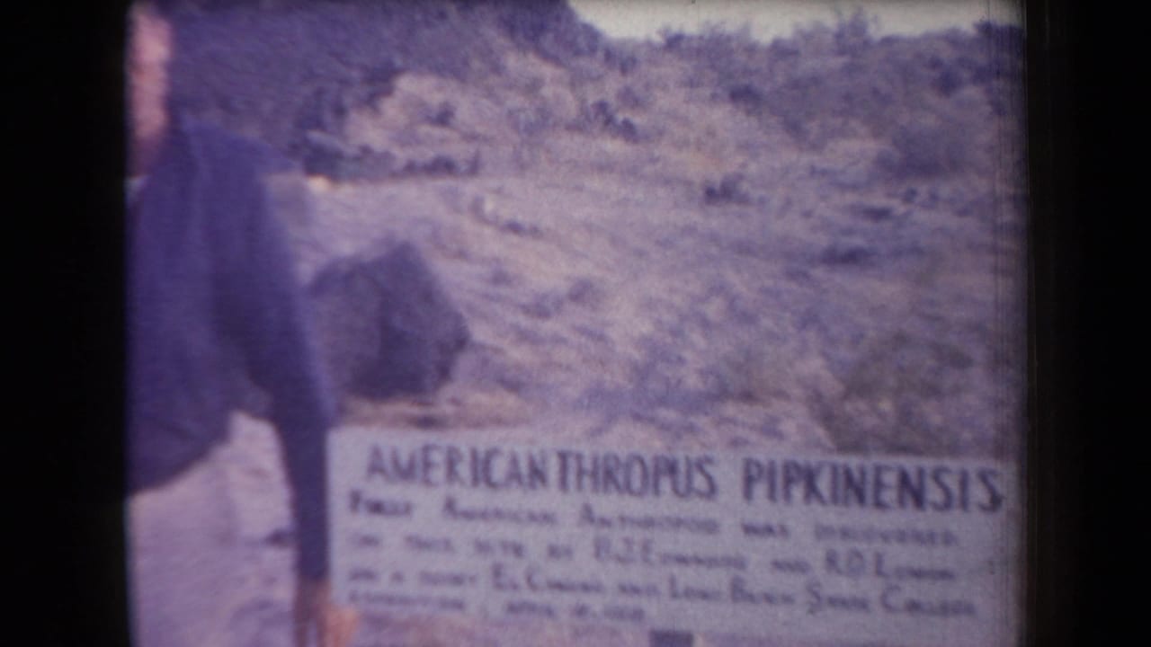 1958 Arizona home-movie frame showing a desert slope, scrub vegetation, and a sign reading AMERICANTHROPUS PIPKINENSIS.