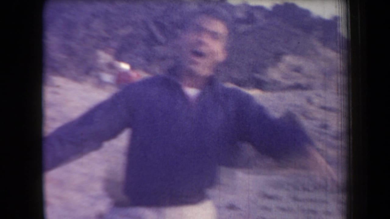 A man in a dark sweater and light pants gestures broadly toward the camera in the Arizona desert, filmed on soft 8mm stock.