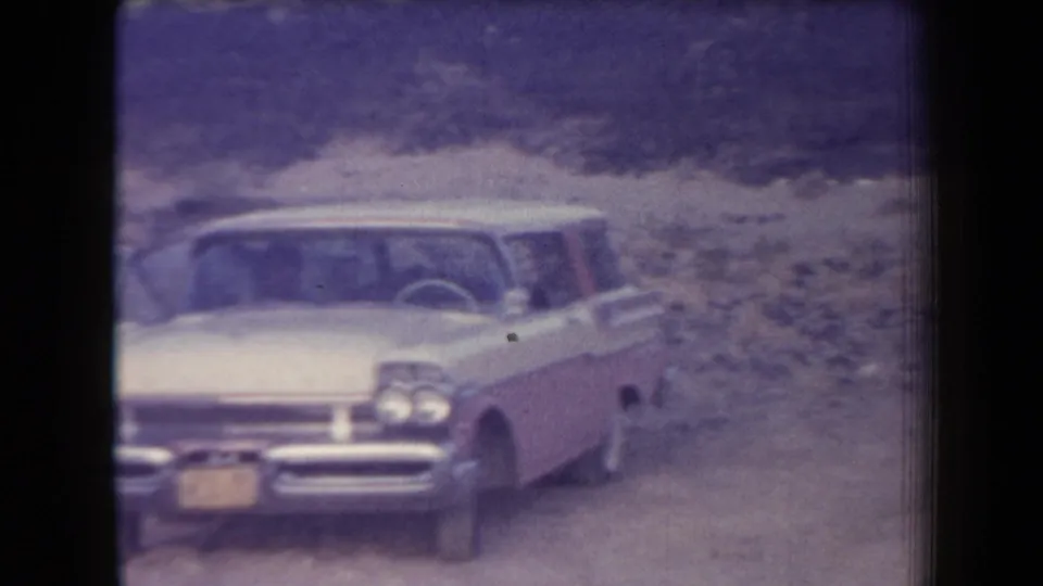 Faded 8mm frame of a 1950s sedan facing the camera on a dusty Arizona desert road.