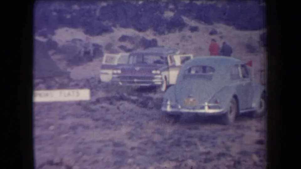 1958 Arizona frame showing a light blue Volkswagen Beetle, other parked cars, and distant people in a rough outdoor parking area.