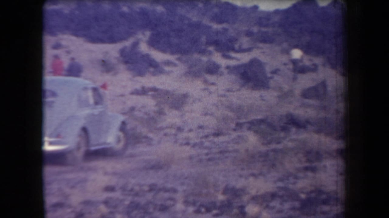 A pale classic car appears near the edge of a rocky Arizona desert scene, with scrub and hills in the background.