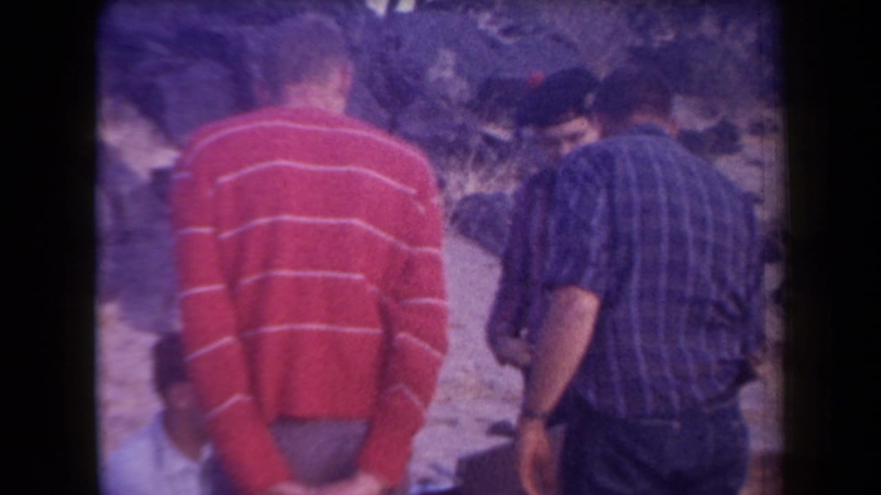 1958 Arizona home-movie frame showing several adult men gathered in a rocky desert setting, seen from behind and partly obscured.