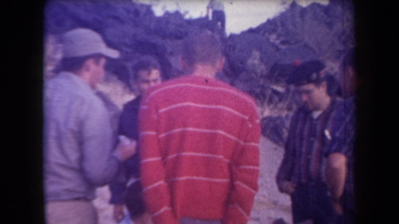 Five men gathered outdoors in desert terrain, including one in a red striped sweater and one in a light cap.
