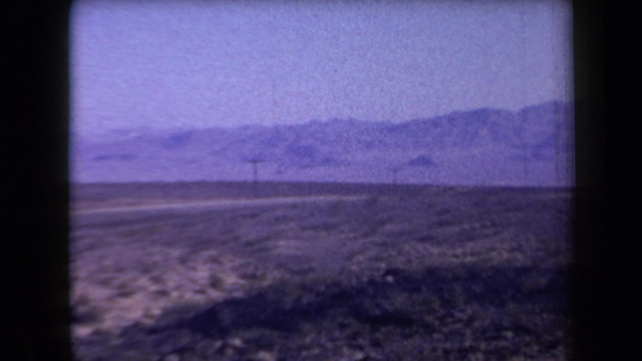 1958 Arizona 8mm frame showing a desert road, sparse shrubs, utility poles, and low mountains in the distance.