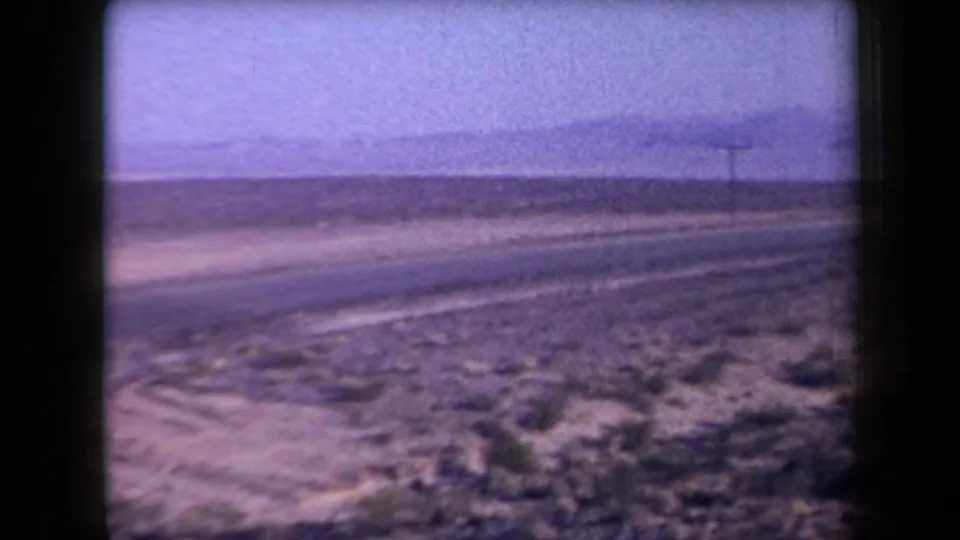 1958 Arizona 8mm frame of a moving roadside view with brushy foreground, utility pole, road edge, and distant mountain ridge.