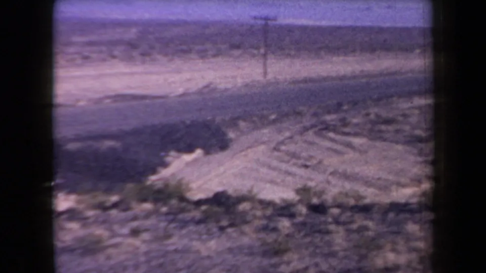 1958 Arizona 8mm frame showing a sandy roadside mound, open desert brush, a utility pole, road surface, and distant hills.