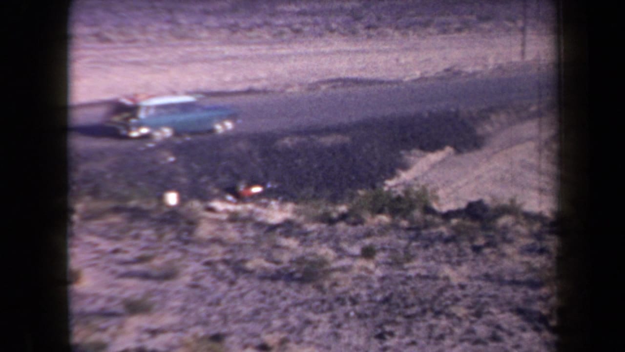 1958 Arizona home-movie frame showing a turquoise station wagon near sandy terrain with sparse scrub and heavy film grain.