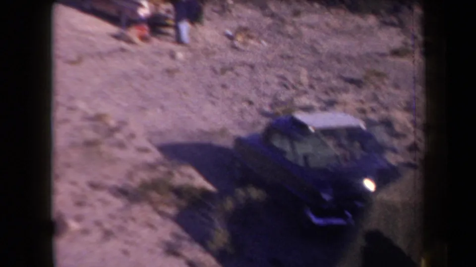 1958 Arizona roadside camping view with a dark blue sedan, a tent edge, and distant people on sandy ground.