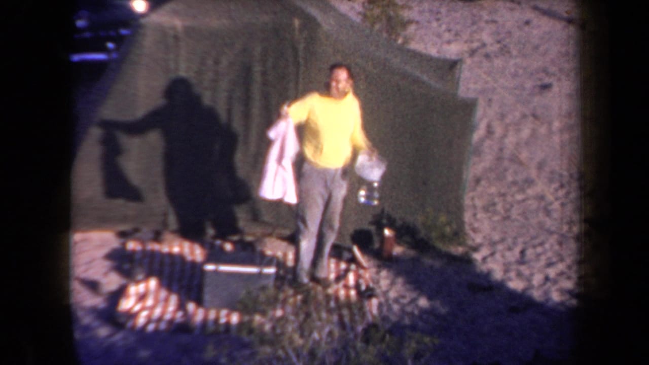 1958 Arizona camping scene with a man holding a pink garment and bottle near a green tent and scattered camp gear.