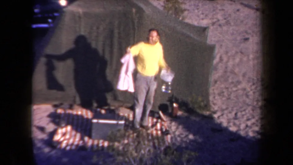 1958 Arizona camping scene with a man holding a pink garment and bottle near a green tent and scattered camp gear.