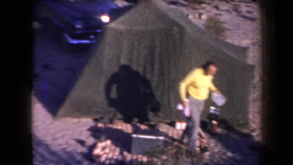 Home-movie frame from a 1958 Arizona campsite showing a man moving beside a canvas tent, blanket, and parked sedan.