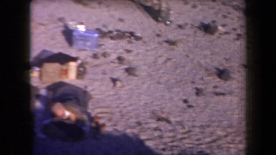 1958 Arizona campsite detail showing sandy ground, a blue cooler, a box, rocks, and partial camp items at frame edge.