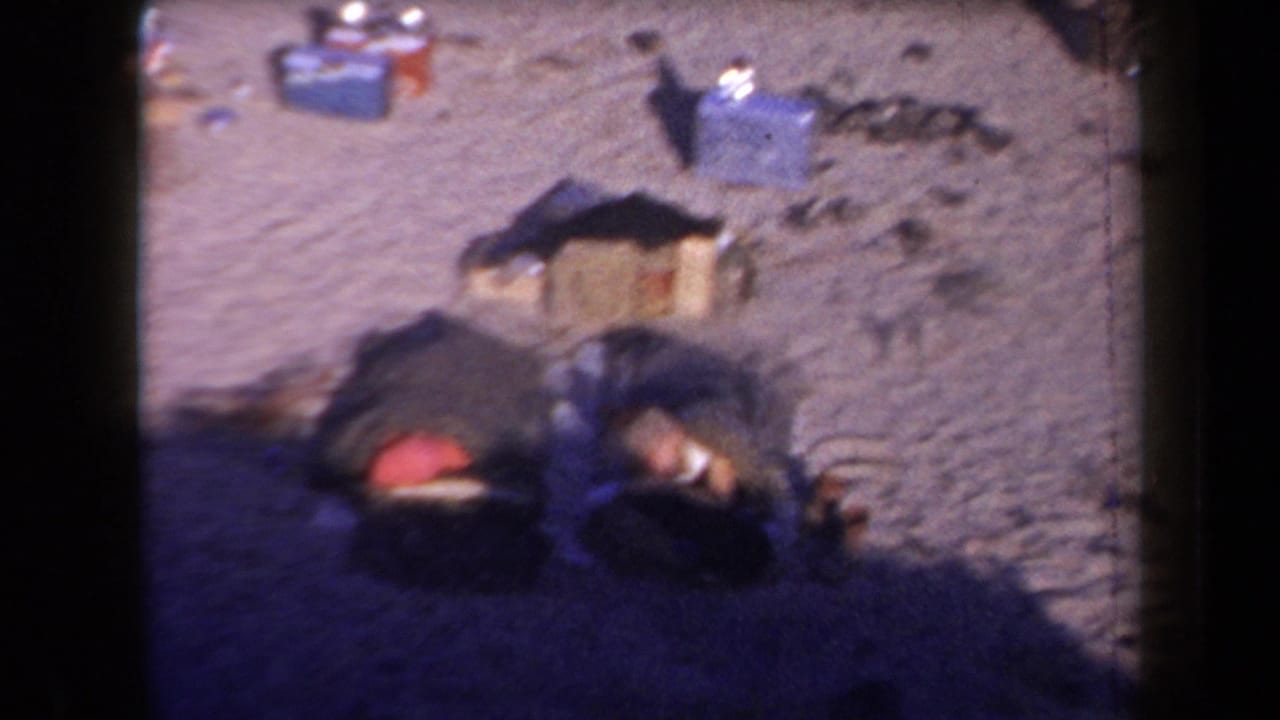 1958 Arizona home-movie frame showing people lying in sleeping bags at a sparse desert campsite with gear nearby.