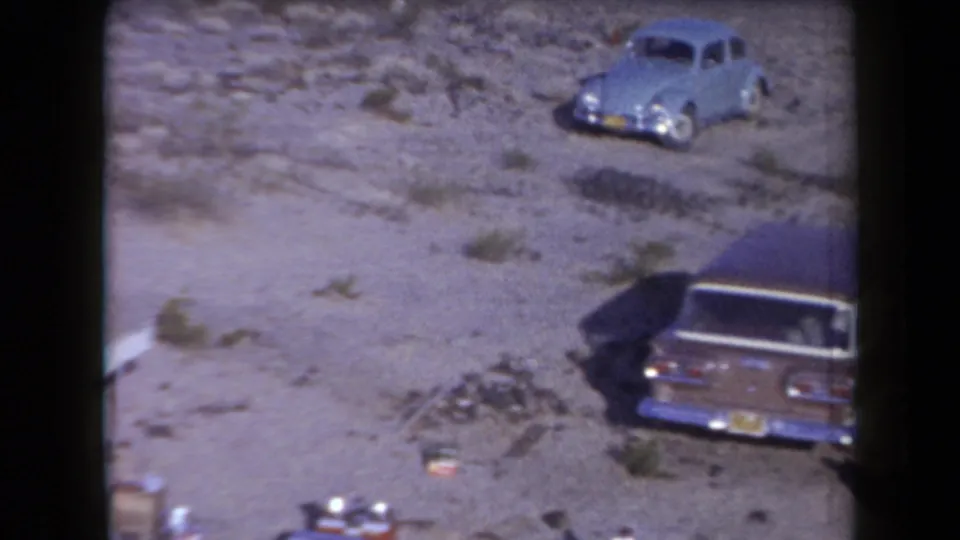 Two mid-century cars parked on open Arizona desert ground, with campsite gear partly visible nearby.