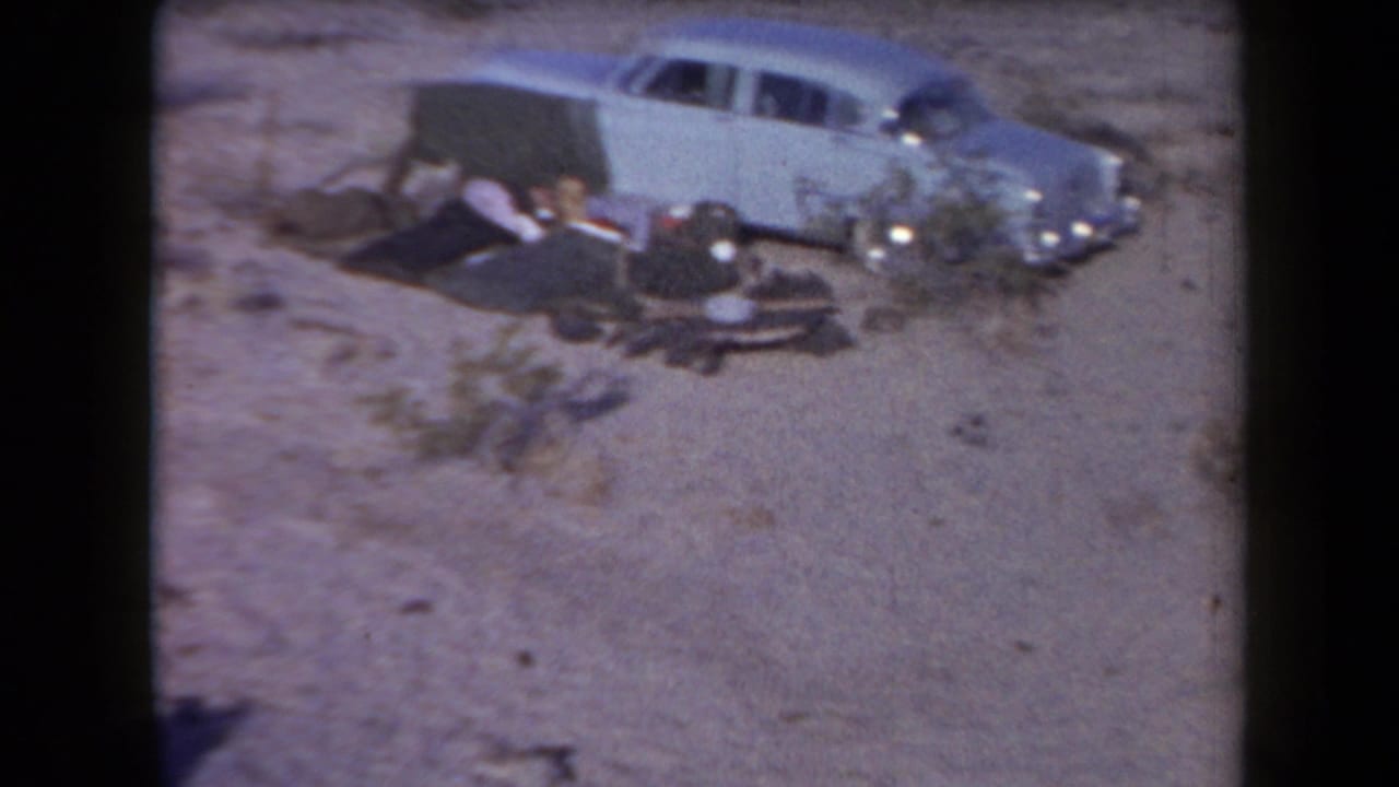 Light-colored sedan overturned in the desert with people and belongings nearby.