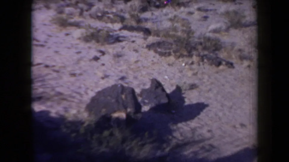 1958 Arizona 8mm frame showing the overturned vehicle from closer range, partly in shadow on sandy desert terrain.