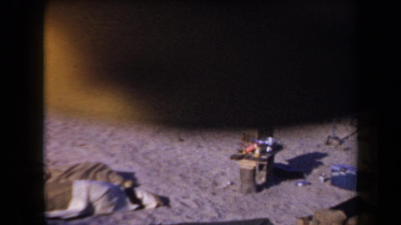 1958 Arizona home-movie frame showing a sandy campsite, a person resting on a blanket, and camp gear under heavy film shadow.