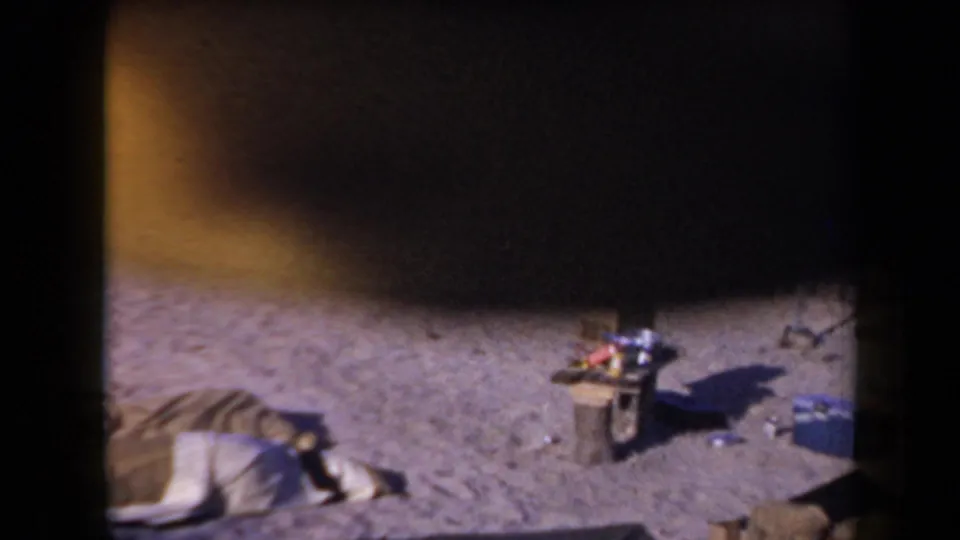 1958 Arizona home-movie frame showing a sandy campsite, a person resting on a blanket, and camp gear under heavy film shadow.