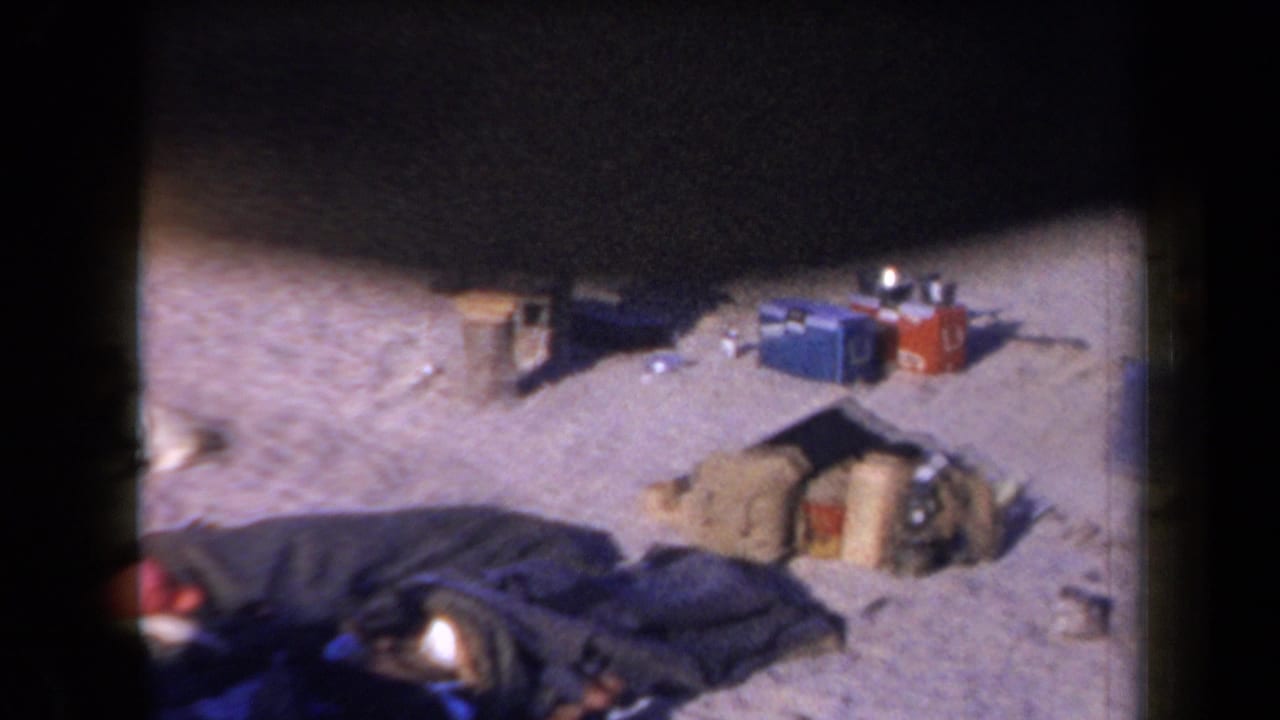 1958 Arizona desert campsite with blankets, bags, coolers, and scattered camp supplies visible in a grainy 8mm frame.