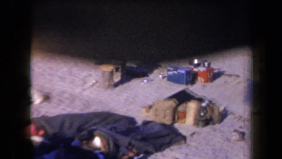 1958 Arizona desert campsite with blankets, bags, coolers, and scattered camp supplies visible in a grainy 8mm frame.