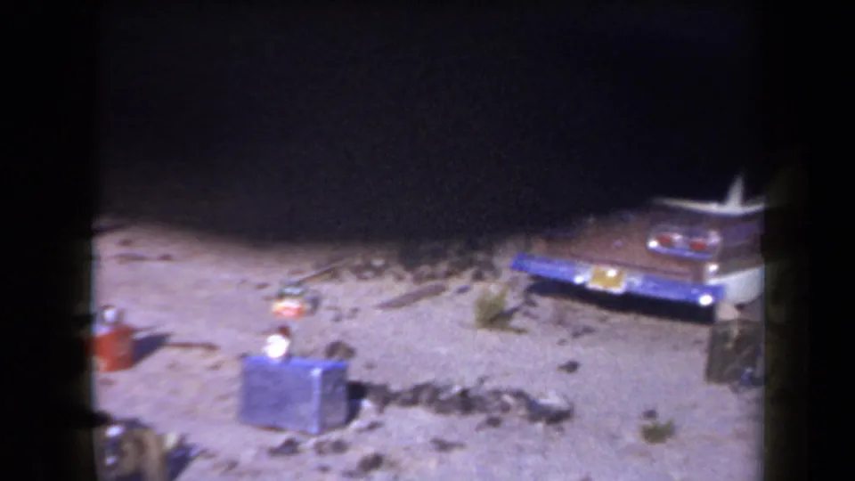 1958 Arizona home-movie frame showing the rear of a station wagon with coolers and camp equipment set out in the sand.