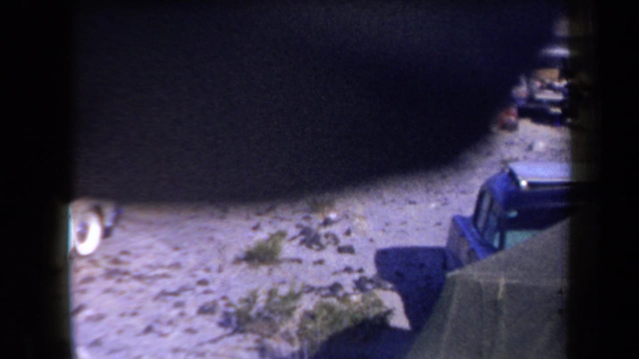 1958 Arizona 8mm frame showing a dim campsite area with a tent edge, sandy ground, and parked cars partly visible beyond the shadows.