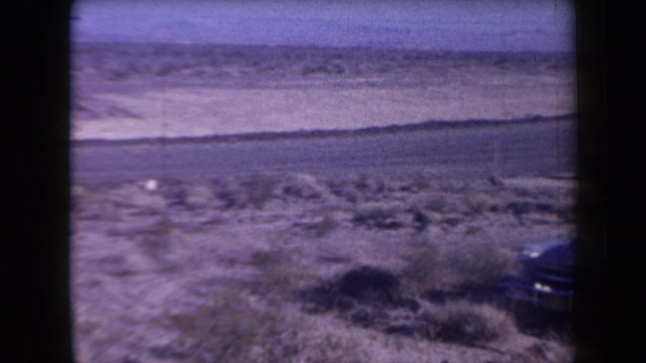 Desert highway and scrubland with part of a parked dark car at the right edge.
