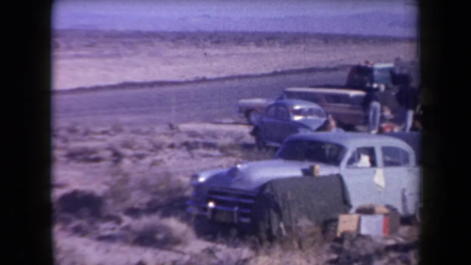 1958 Arizona home-movie frame showing several 1950s cars parked in desert scrub, with people resting near luggage or boxes.