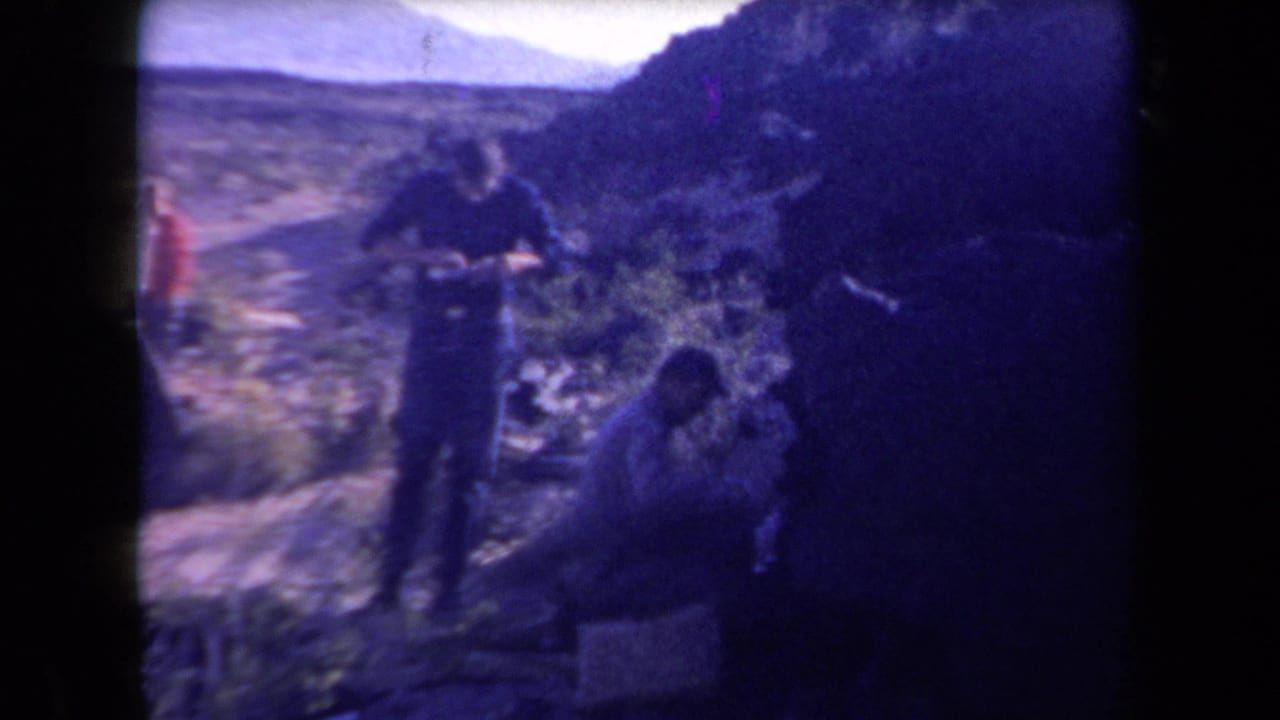 1958 Arizona home-movie frame showing men in a rugged desert area, with one standing and another crouched near brush and rocks.