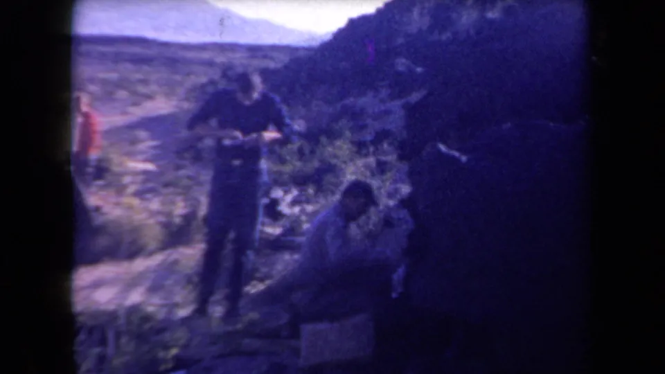 1958 Arizona home-movie frame showing men in a rugged desert area, with one standing and another crouched near brush and rocks.