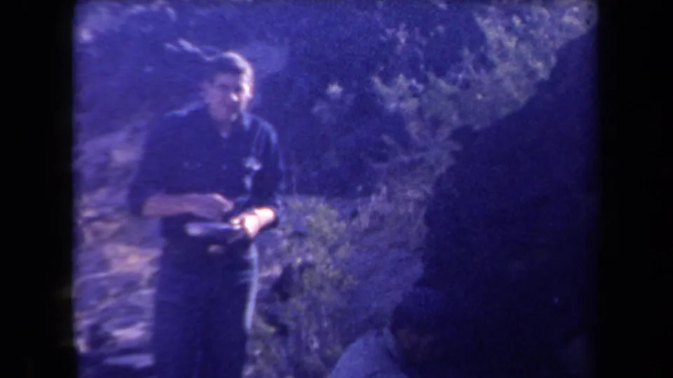 Adult man in 1958 Arizona home-movie footage stands in desert terrain holding a plate or dish, with heavy purple color cast.