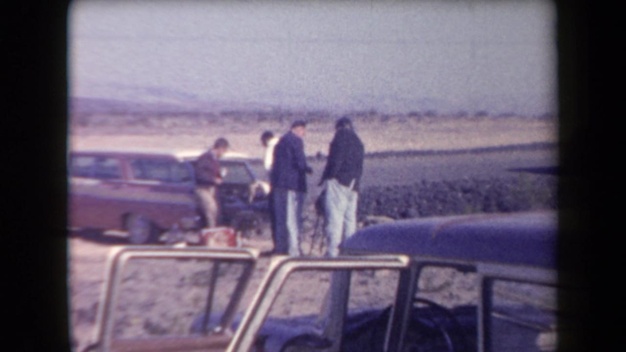 1958 Arizona home-movie frame showing several men standing near a red station wagon on a desert roadside.