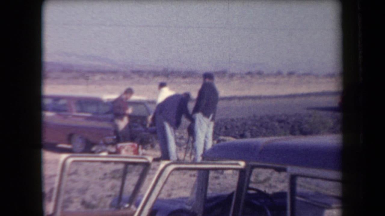 1958 Arizona frame with men inspecting or handling gear near a red station wagon in an open desert landscape.