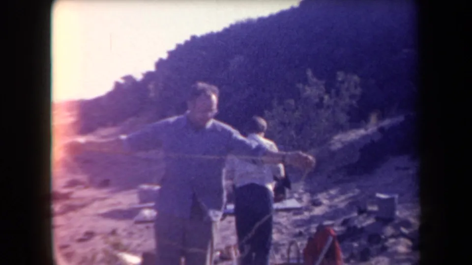 1958 Arizona home-movie frame showing a man gesturing near brushy hills, with another person and gear in the background.