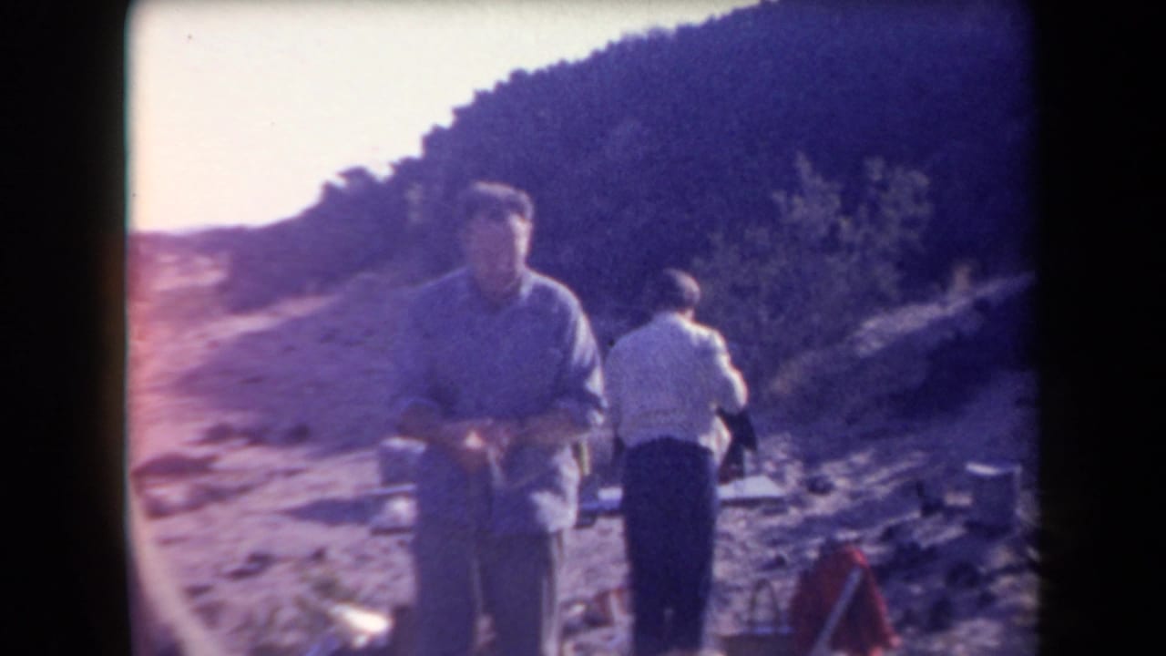 1958 Arizona 8mm frame showing two adult men near camp gear on rocky desert ground, with a hillside and scrub in the background.