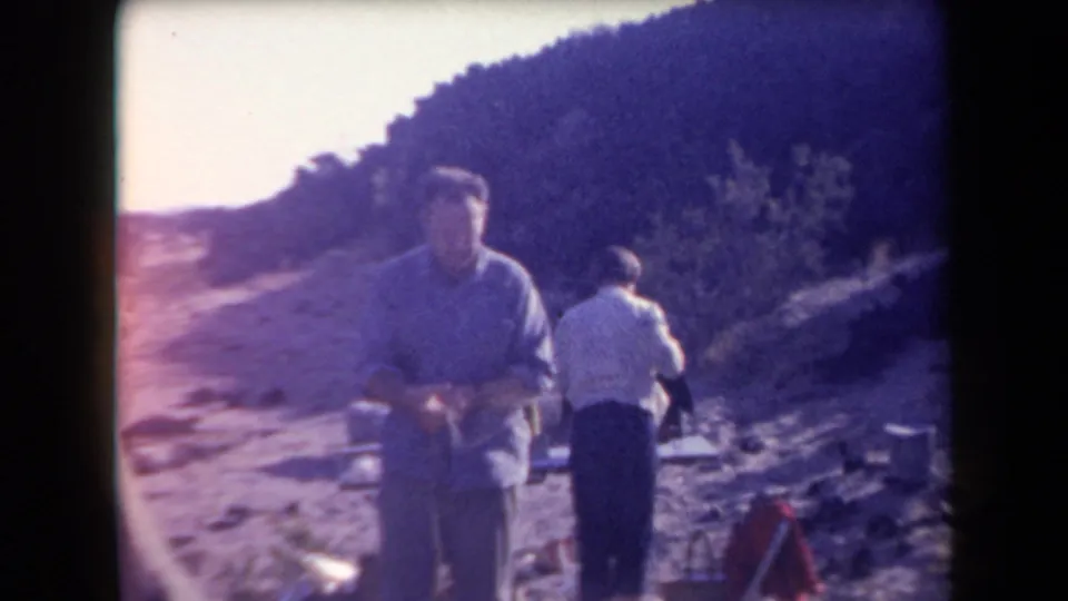 1958 Arizona 8mm frame showing two adult men near camp gear on rocky desert ground, with a hillside and scrub in the background.